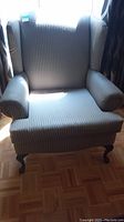 Front view of wing back chair with padded striped upholstery and visible wooden cabriole legs, sitting on parquet floor, sunlight casting shadows.