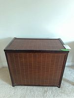 Front view of the rectangular rattan and wood laundry hamper placed against a wall on carpeted floor.
