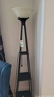 Full front view of a black metal floor lamp with three triangular shelves and a white frosted bowl shade at the top, situated against a wall next to a blue fabric chair.