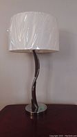 Single table lamp with curved metal base and white drum-shaped shade wrapped in plastic, positioned on a dark wood surface against a light wall.