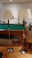 Two floor lamps standing in front of a pool table, showing overall appearance, bases, wooden midsections, and fabric lampshades.