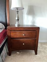 Front view of a wooden nightstand with a lamp on top, showing two drawers with metal knobs.