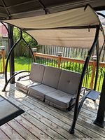 Full view of the black metal frame canopied swing chair with beige fabric canopy and three gray seat cushions arranged upright, showing overall wear and condition.