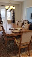 Full view of wood pedestal dining table with leaf and five upholstered chairs on each side, placed on patterned carpet with light fixture above.