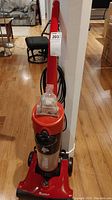 Front view of red Dirt Devil Clean Vision upright vacuum with clear dirt collection cup, black power cord, and handle.