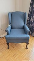 Front view of the blue upholstered wing chair showing the curved wooden legs and cushioned seat.