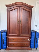 Full front view of cherry wood wardrobe with two doors and two bottom drawers, flanked by blue suitcases for scale