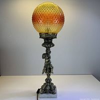 Full view of table lamp illuminated, showing amber hobnail glass globe, cherub figure, and marble base.