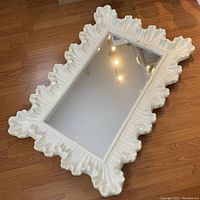 Large decorative mirror lying flat on wooden floor, showing full frame and mirror surface.