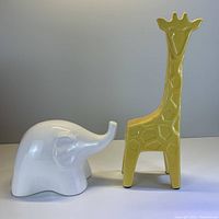 Front and side views of yellow giraffe and white elephant ceramic figurines on white surface with neutral gray background