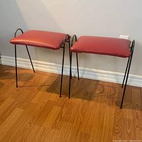 Pair of vintage mid-century stools showing red vinyl cushioned seats and slender black metal legs, set against a wooden floor and white wall.