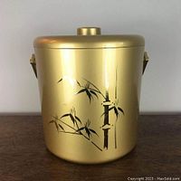 Front view of retro gold-colored ice bucket with black bamboo design and lid, on wooden surface.