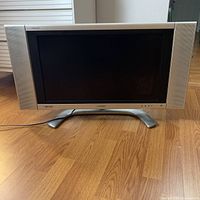 Front view of Sharp Aquos flat screen TV powered off showing silver frame and integrated speakers on sides with pedestal stand on wooden floor.