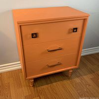 Front view of vintage smallboy dresser with three drawers, painted in warm orange color, showing all handles and legs.