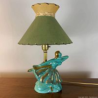 Full view of vintage mid century ceramic ballet dancer table lamp with green and beige fabric shade on wooden surface.