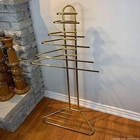 Front view of vintage gold-tone floor tower stand showing tiered horizontal rods and triangular base on wood floor near fireplace.