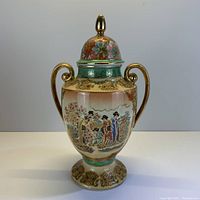 Vintage Satsuma ginger jar with intricate hand-painted traditional Japanese figures, two gold handles, and lid with finial, photographed against gray background.