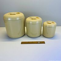 Front view of three beige ceramic canisters in graduated sizes with lids on white background, measuring ruler placed in front for scale.
