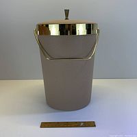 Front view of vintage tall ice bucket showing taupe textured exterior, gold rim, and gold handle, with ruler at base for scale.