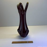 Front view of vintage stretch art glass vase with ruler for scale, showing three upward-flaring elongated arms and deep reddish-purple color.