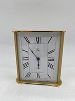 Front view of brass desk clock showing white face with black Roman numerals, brand name Ergo, black hands, and brass frame.