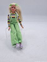 Full body image showing the doll seated, wearing green overalls, striped shirt and purple shoes.