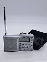 Full front view of the Grundig portable radio with antenna extended and carrying case beside it, showing overall design and layout.