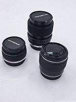 Three Olympus lenses are displayed on a white surface with front lens caps on showing the Olympus brand.