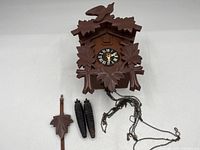 Wooden cuckoo clock with two pine cone-shaped weights, leaf-shaped pendulum and chains shown on surface