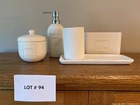 5 piece bathroom vanity set including lotion jar, lotion pump, toothbrush holder, soap dish, and organizer container. All white ceramic with embossed labeling.