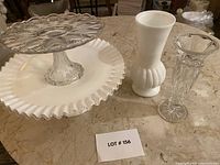 All four items shown together on a marble surface: two cake stands (clear feathered glass and white ruffle edge glass) and two vases (taller white and smaller clear bud vase).