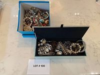 Two boxes are shown, one open blue box filled with assorted costume jewelry, and one open modern jewelry box filled with various jewelry pieces such as bracelets, rings, and earrings.