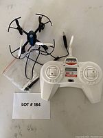 Photo of the Holy Stone drone with controller and accessories on a light surface. The drone is black and white, the controller is white with joysticks and buttons. Spare propellers and a charging cable included.
