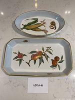 Top view of two ceramic serving dishes: one oblong with peach design, one oval with corn design, both with gold trim