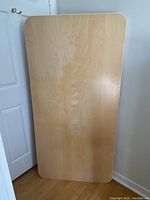 Full view of the rectangular Ikea tabletop standing upright against a wall, showing smooth light wood grain surface with rounded corners.