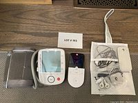 Two Life Brand medical devices and related accessories spread on the floor including a blood pressure monitor, its cuff, a pulse oximeter with remote, and a mesh pouch with wires and clips.
