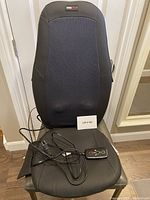 Front view of the black massage cushion placed on a chair. Wired remote with buttons for massage operation is resting on the seat.