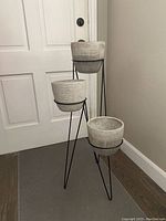 Full view of the three-tiered black metal plant stand with three light gray ceramic pots placed indoors against white wall and door, showing overall size and design.