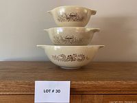 Three vintage Pyrex Cinderella nesting bowls stacked, showing brown floral and nature themed patterns against tan and cream backgrounds.