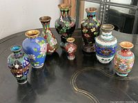 Nine hand-painted cloisonne vases shown together on a black table, displaying diverse floral designs and colors.