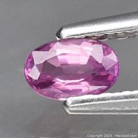 Close-up image of an oval cut natural sapphire gemstone, pink-purple in color, held by tweezers showing facets and clarity.
