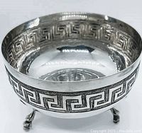 Close-up side angle of the circular silver bowl showing geometric Greek key pattern, claw feet, and polished silver finish.