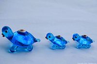 Three blue glass turtle figurines shown side by side, varying sizes large, medium, and small. Each has prominent eye detail on the shell and red mouth detail.