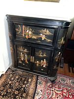 Front view of Asian black lacquered bar cabinet showing relief design with figures and landscape scenes on drawer and doors.