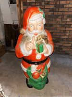 Full front view of the lighted blow mold Santa figure placed indoors against a brick wall, showing details of Santa's face, red suit, and green sack with toys.