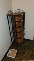 Corner shelf constructed of black metal frame with wooden top and four wicker basket drawers. Bathroom scale on the floor nearby.