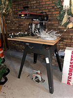 Craftsman 10-Inch Radial Arm Saw mounted on a black metal stand with wooden tabletop in a basement environment, showing the full setup from the side including surrounding decor and packaging boxes.