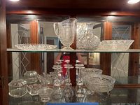 Glass cabinet with crystal and glass items including wine glasses, decanter, bowls, covered box, creamer, and sugar bowl on glass shelves.