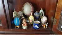 Front view of the collection including decorative beaded eggs in various colors and styles, a blue cup, Goebel Hummel boy figurine, and small colorful figurines.