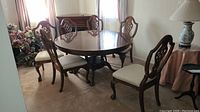Six vintage wooden dining chairs arranged around a round dining table in room with carpet. Chairs have carved backs and beige upholstered seats.